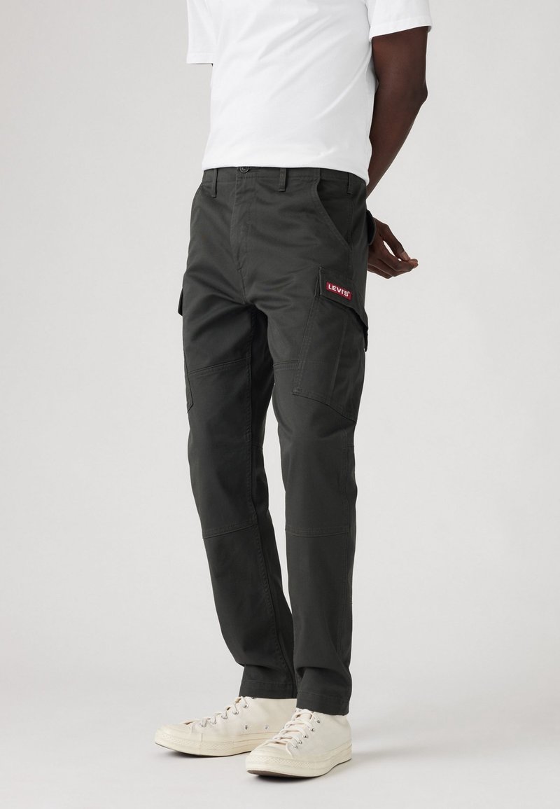 Personne portant un pantalon cargo noir avec le logo Levi's sur la poche, des baskets blanches et un t-shirt blanc sur un fond clair uni.
