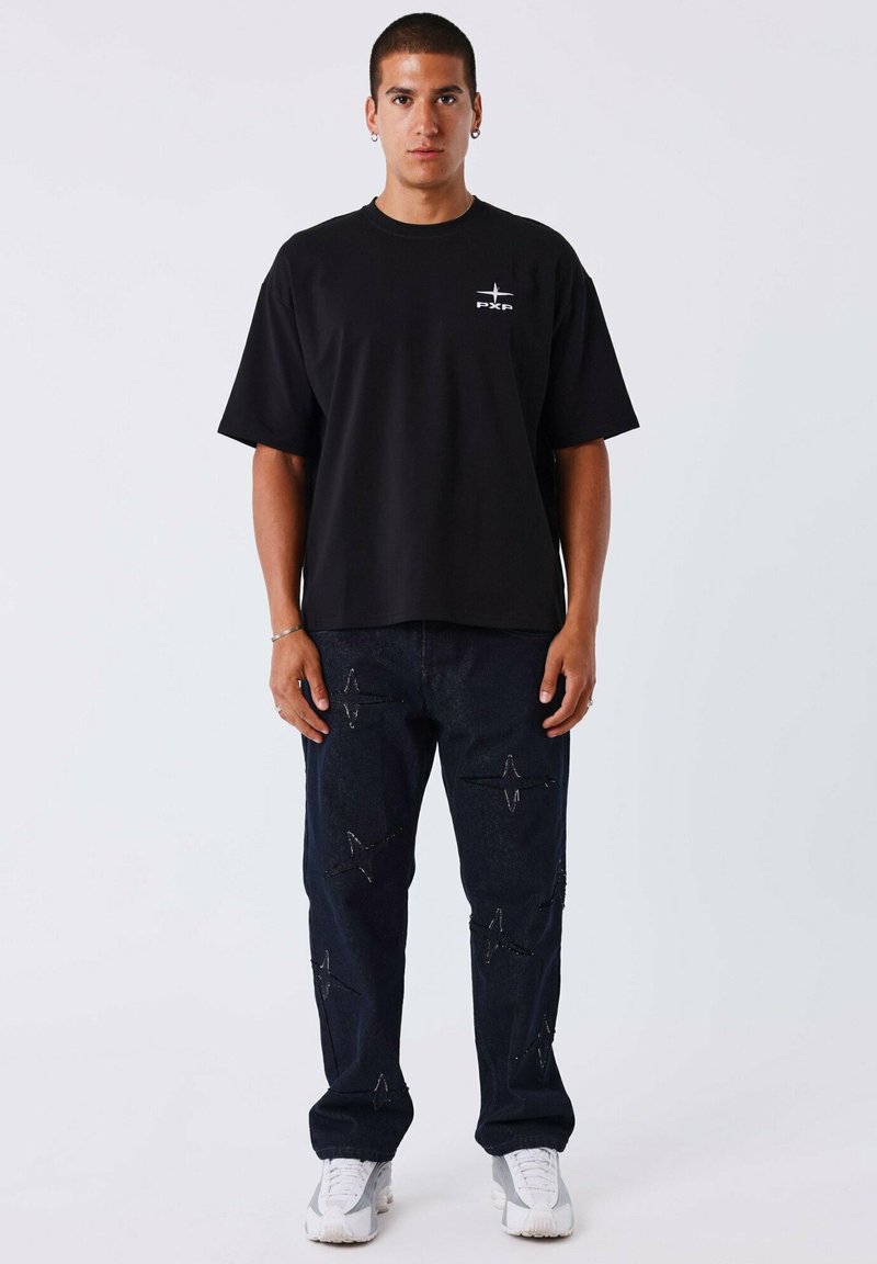 Camiseta negra de gran tamaño combinada con pantalones de mezclilla oscuros que presentan patrones en forma de estrella. Llevada con zapatillas blancas, creando un conjunto casual.