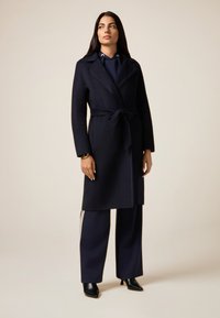 Cappotto in misto lana blu navy con vita regolabile, colletto a bavero e dettagli cuciti, abbinato a pantaloni larghi blu navy e tacchi neri a punta.