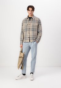 Checkered beige and navy flannel shirt, buttoned, worn over a black top; light blue trousers; white sneakers; holding a beige bag.