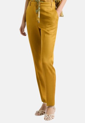 Donna con pantaloni su misura color senape e sandali beige con cinturini, mani in tasca, su sfondo uniforme.