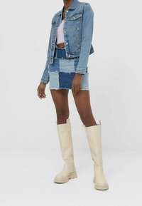 Spijkerjack met lichtblauwe en middenblauwe patchwork minirok. Beige kniehooglaarzen met een dikke zool. Heldere achtergrond.