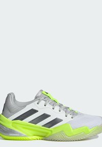 Zapatilla deportiva blanca con acentos grises y destacados en amarillo-verde brillante. Parte superior de malla, suela texturizada y tres rayas grises en el lateral.