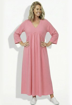 Lächelnde Frau mit blondem Haar steht mit Händen in der Hüfte, trägt ein langes rosa Kleid und weiße Turnschuhe vor einem schlichten weißen Hintergrund.