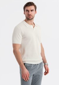 Stickad kortärmad polo i off-white med ribbat krage och ärmslut, med ett texturerat mönster. Tillsammans med ljusgrå randiga byxor.