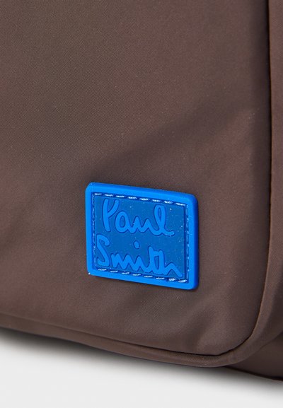 Pruun nailonist kott, mille esiküljel on sinine ristkülikukujuline logo silt, millel on tõstetud tekst "Paul Smith". Sile tekstuur, kompaktne disain.