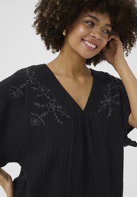 Fekete tunika V-nyakkivágással, amely tonálisan hímzett virágmintával díszíti a vállakat, valamint texturált anyagával és pliszírozott kialakításával rendelkezik.