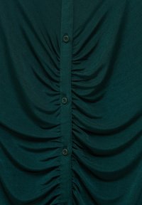 Blouse turquoise avec texture froncée, dotée d'une rangée de boutons sombres sur le devant, confectionnée dans un tissu lisse et léger.