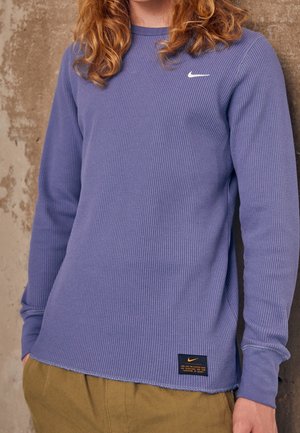 Junge Person trägt ein lila strukturiertes langärmeliges Nike-Shirt und beige Hose, steht vor einer abgenutzten Betonwand.