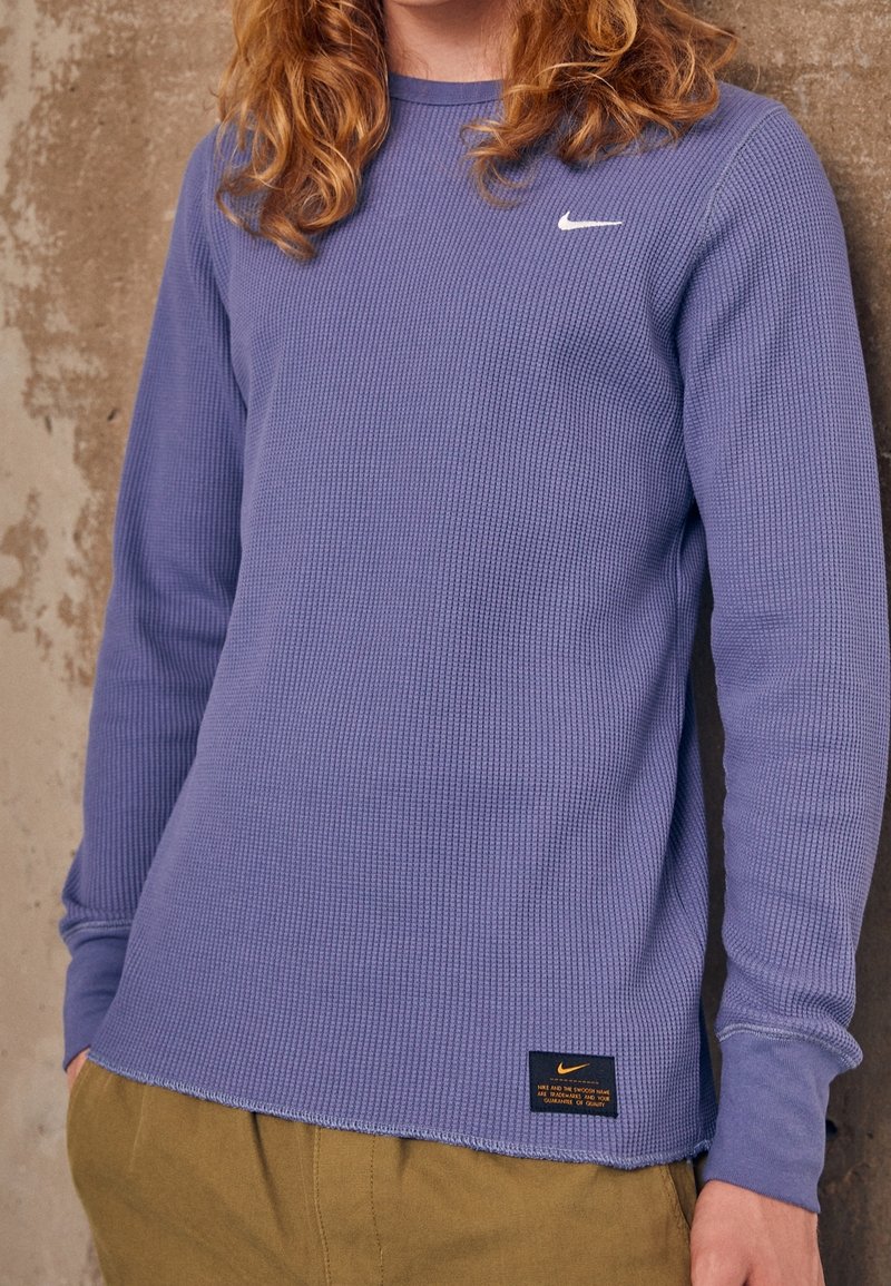 Jeune personne portant un t-shirt Nike violet texturé à manches longues et un pantalon beige, debout contre un mur en béton usé.