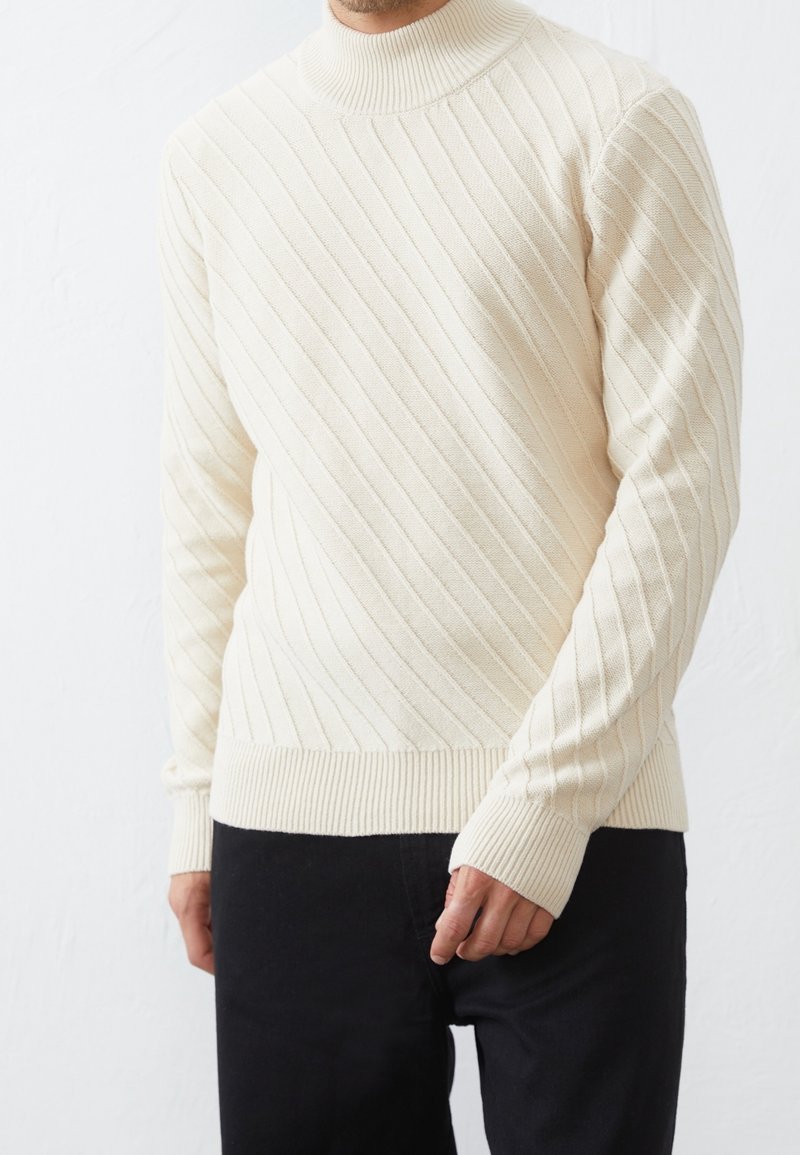 Man klädd i en krämfärgad ribbad stickad polotröja och svarta byxor, stående mot en enfärgad vit bakgrund.