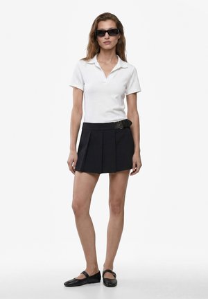 Mujer de pie con gafas de sol negras, camisa blanca de manga corta con cuello, minifalda negra plisada con cinturón y zapatos planos negros.