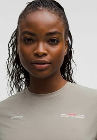 Nahaufnahme einer Frau mit geflochtenem Haar, die ein hellgraues Lululemon-Shirt mit dem Text "Finde den Fluss auf der Reise." trägt.