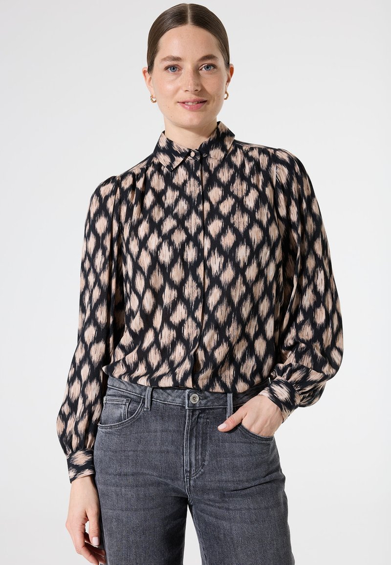 Blouse à motifs noir et beige avec un col haut, des manches longues et une fermeture par boutons, associée à un jean gris. Le tissu semble léger.