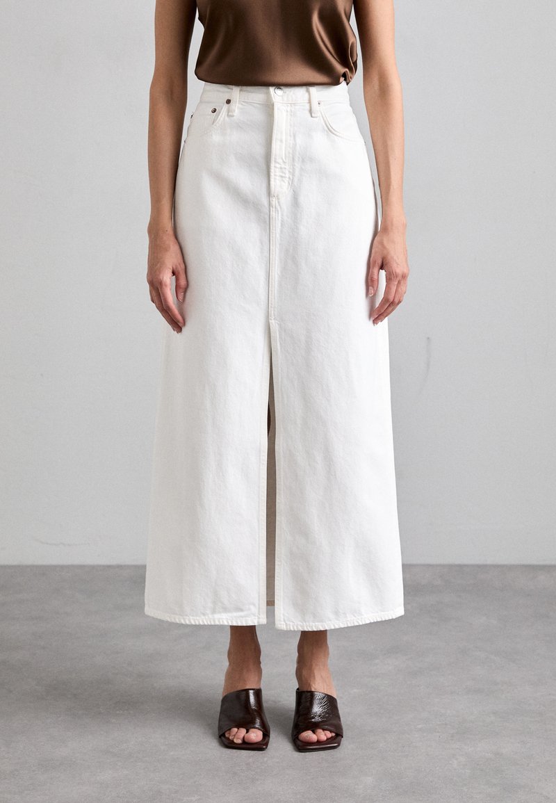 White denim maxi skirt with front slit, high waist, and tonal stitching. Paired with dark brown open-toed mules.