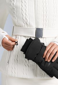 Giacca bianca con trama a trecce, con cerniera argentata e un guanto nero tenuto in mano. Fibbia argentata su una cintura larga.