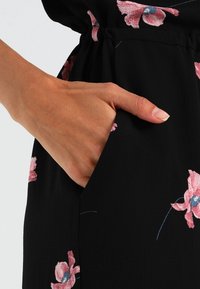 Tejido negro con un patrón floral en tonos rosa y azul, mostrando una mano colocada en un bolsillo lateral. Textura suave, diseño ligero.