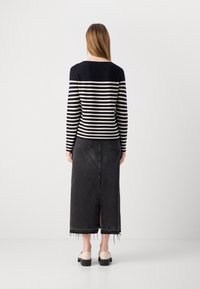 Kobieta stoi tyłem, ubrana w czarno-białą, prążkowaną bluzkę z długim rękawem, czarną, postrzępioną spódnicę dżinsową i beżowe buty.