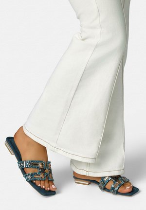 Stopy w białych dzwonowatych spodniach i niebieskich sandałach z odkrytymi palcami, ozdobionych paskami z biżuterią i niskimi kwadratowymi obcasami na białym tle.