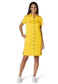 Kurzärmliges gelbes Kleid mit Kragen, zwei Brusttaschen, durchgehender Knopfleiste und Schlitzen an den Seiten. Styliert mit weißen Sneakers.
