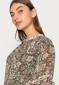 Blusa de estampado floral em verde oliva, com flores rosa e brancas, um decote redondo e detalhes de mangas com babados. Tecido leve.