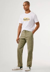 Hvit T-skjorte med blått og gult logo, kombinert med lyse olivengrønne bukser. Modellen har på seg hvite joggesko med grønne detaljer.