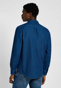 Granatowa koszula dżinsowa z długimi rękawami, z guzikami przy mankietach, spiczastym kołnierzem i plisą na plecach. Tkanina ma gładką fakturę.