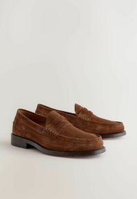 Mocasines de ante marrón con un diseño cosido, punta redonda y un tacón bajo negro. La parte superior presenta un acento elevado a lo largo de la costura.