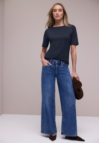 Kvinde stående med den ene hånd i lommen, iført en sort top med korte ærmer, blå bukser med vidt ben, brune spidse sko og holder en brun hat.