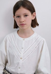 Blusa blanca de algodón con cuello redondo, presenta cortes verticales, cierre de botones y mangas largas. Diseño simple y transpirable.