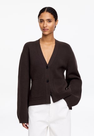 Woman wearing a dark brown buttoned cardigan with two front pockets and white pants, standing against a plain white background.