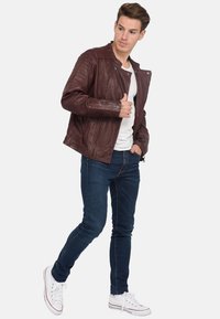 Burgunderfarbene Lederjacke mit vertikalen Nähdetails, Reißverschlusstaschen und Druckknopfkragen, kombiniert mit schmal geschnittenen blauen Jeans und weißen Turnschuhen.