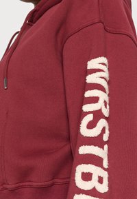 Viininpunainen huppari, jossa on pehmeä beige logo hihassa, kiristysnauhahihoissa ja resorit hihansuissa. Pehmeä materiaali ja rento istuvuus.