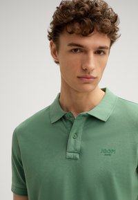Polo verde realizzato in tessuto testurizzato. Caratterizzato da un colletto classico, un'apertura con due bottoni e il logo ricamato "JOOP! JEANS" sul petto.