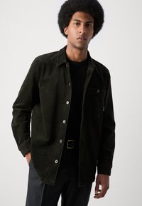 Camisa de corduroy verde oscuro con botones, un bolsillo en el pecho y un corte relajado. Llevada sobre una camiseta negra. Estilizada con pantalones negros.