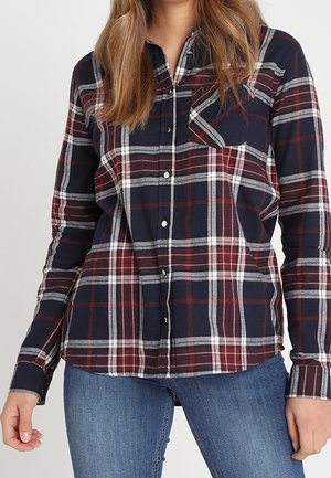 Kobieta ubrana w granatową, czerwoną i białą koszulę w kratę zapinaną na guziki z kieszenią na piersi oraz niebieskie dżinsy, na tle jednolitego jasnego tła.