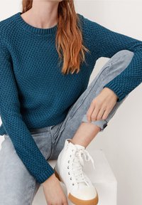 Turkusowy sweter z dzianiny o teksturowanym wzorze, zestawiony z jasnoszarymi jeansami z podwiniętymi nogawkami oraz białymi sneakersami na wysokiej cholewce z gumową podeszwą.