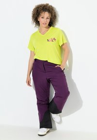 Kvinde med krøllet hår iført en lys gul V-hals T-shirt og lilla skibukser, stående med den ene hånd i lommen mod en hvid væg.