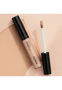 Concealer mit voller Deckkraft in einem transparenten Röhrchen mit schwarzem Deckel. Der Applikator ist ein weißer Stab; seine Spitze zeigt eine hellbeige Cremetextur.