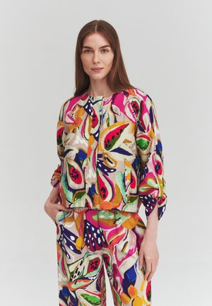 Frau mit langen braunen Haaren trägt eine passende bunte Blumenjacke und Hose, die linke Hand in der Tasche, vor einem einfarbigen Hintergrund.
