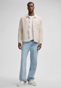 Giacca beige testurizzata con bottoni frontali, tasca singola sul petto, indossata sopra una maglietta bianca e jeans azzurri larghi, abbinata a sneaker bianche.