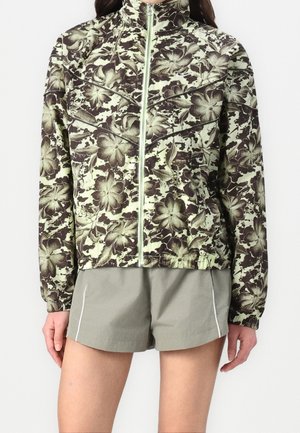 Groen en bruin bloemenpatroon jas met ritssluiting en hoge kraag, gedragen met lichtgrijze shorts met witte zijstrepen.