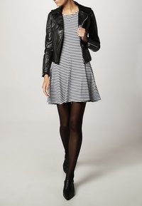 Czarna skórzana kurtka motocyklowa noszona na białej i granatowej sukience w paski, w połączeniu z czarnymi rajstopami i czarnymi czółenkami.