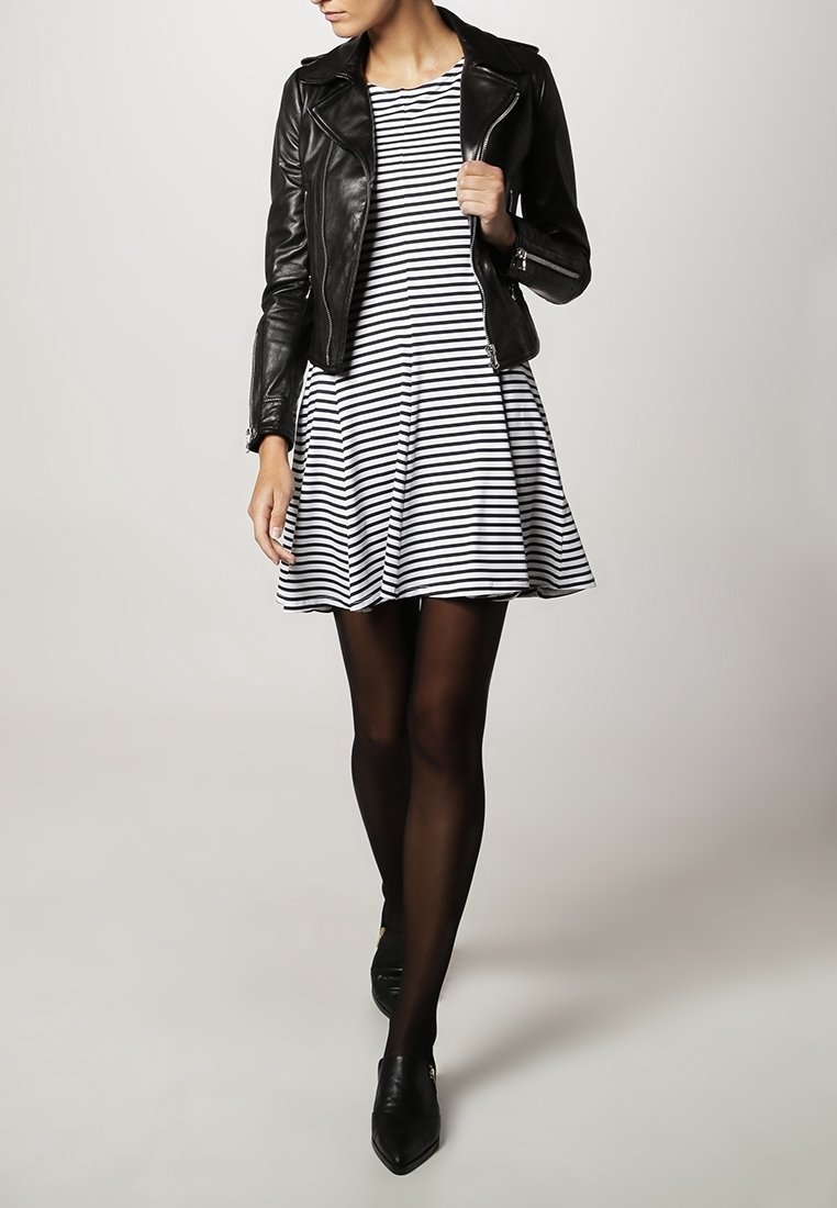 Czarna skórzana kurtka motocyklowa noszona na białej i granatowej sukience w paski, w połączeniu z czarnymi rajstopami i czarnymi czółenkami.