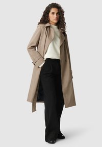 Braune und cremefarbene karierte Trenchcoat mit tailliertem Gürtel, getragen über einem weißen strukturierten Oberteil und schwarzen weiter geschnittenen Hosen.