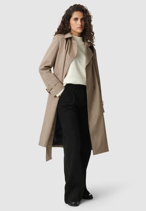 Braune und cremefarbene karierte Trenchcoat mit tailliertem Gürtel, getragen über einem weißen strukturierten Oberteil und schwarzen weiter geschnittenen Hosen.