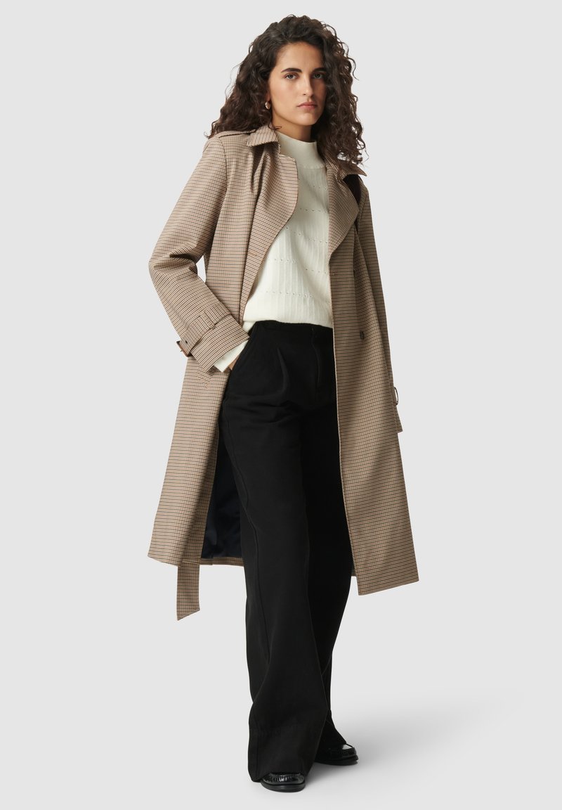 Braune und cremefarbene karierte Trenchcoat mit tailliertem Gürtel, getragen über einem weißen strukturierten Oberteil und schwarzen weiter geschnittenen Hosen.