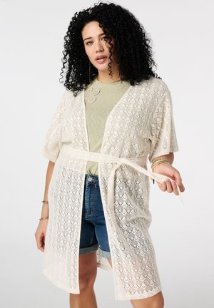 Vrouw met krullend zwart haar, gekleed in een beige kanten vestje met een strik in de taille, over een groen shirt en denim shorts, poserend tegen een effen achtergrond.