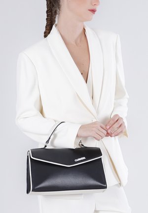 Femme en blazer blanc tenant un sac à main en cuir noir avec bordure blanche et logo argenté, debout devant un fond gris uni.