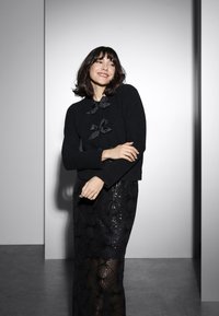 Pull en tricot noir avec trois accents de nœuds en satin, associé à une jupe longue noire à motif géométrique en sequins. Fond mat.